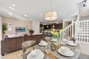 A dining area with a glass table set for six, a modern pendant light, and a kitchen in the background. - Windsor at Westside - 25863 (Kissimmee)