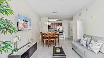 A living room with a sofa, coffee table, and dining area with chairs. There is a kitchen in the background.