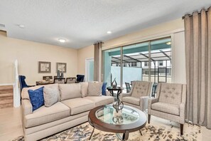 A living room with a beige sofa, armchairs, a glass coffee table, and a large window with curtains. - Windsor at Westside - 26139 (Kissimmee)