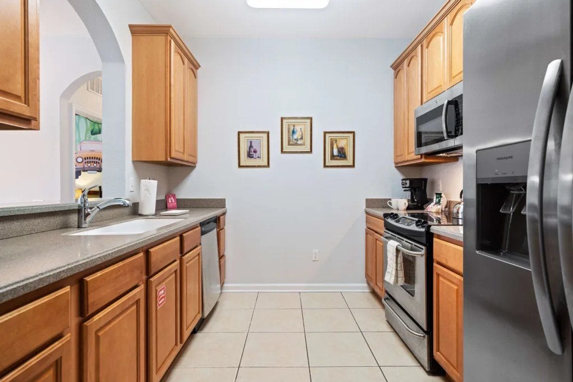 A kitchen with wooden cabinets, a stainless steel refrigerator, a microwave, and a dishwasher. There are three framed pictures on the wall.