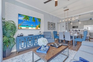 A modern living room with a beach-themed wall art, a blue console table, and a dining area with a kitchen in the background. - Vista Cay - 27376 (Orlando)