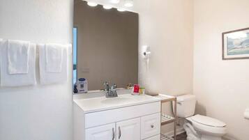 Un baño con lavamanos blanco, espejo y inodoro. Hay toallas colgadas en la pared y un cuadro enmarcado sobre el inodoro.