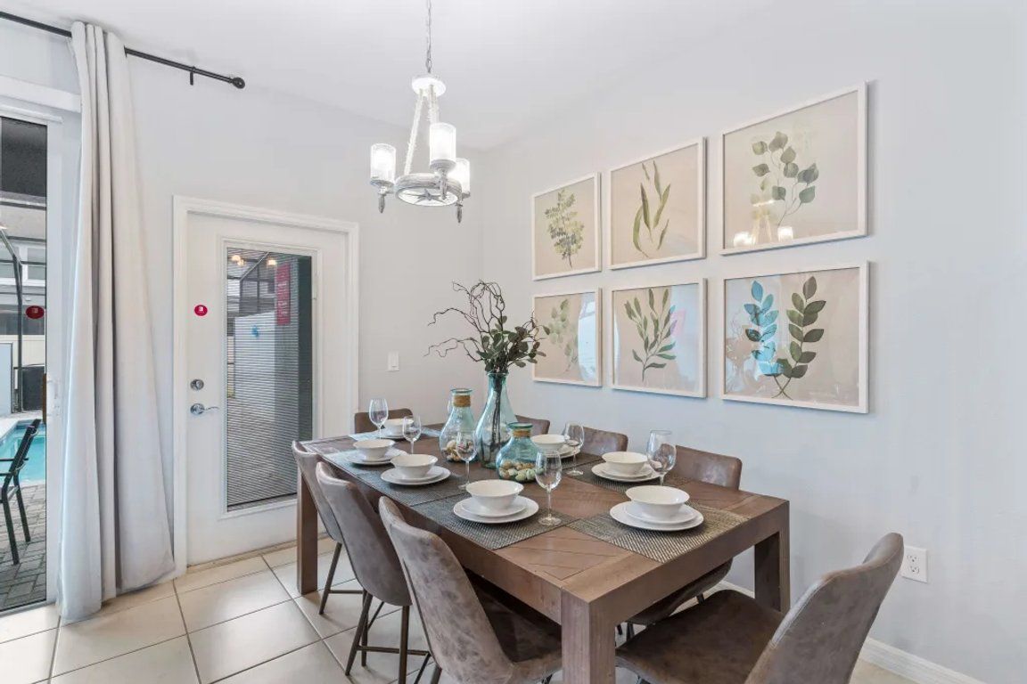 Un comedor con una mesa de madera dispuesta para seis personas, rodeada de sillas. Hay impresiones botánicas enmarcadas en la pared, un candelabro sobre la mesa y una puerta con vistas a una zona de piscina.