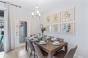 Un comedor con una mesa de madera dispuesta para seis personas, rodeada de sillas. Hay impresiones botánicas enmarcadas en la pared, un candelabro sobre la mesa y una puerta con vistas a una zona de piscina.