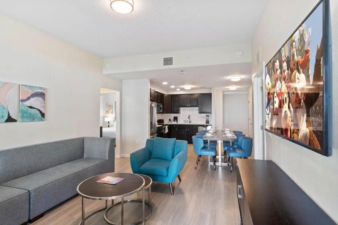 A modern living room with a grey sofa, a round coffee table, and a blue armchair. There is a dining area with a white table and blue chairs. The kitchen is visible in the background with dark cabinets and a white countertop.