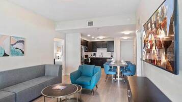 A modern living room with a grey sofa, a round coffee table, and a blue armchair. There is a dining area with a white table and blue chairs. The kitchen is visible in the background with dark cabinets and a white countertop.