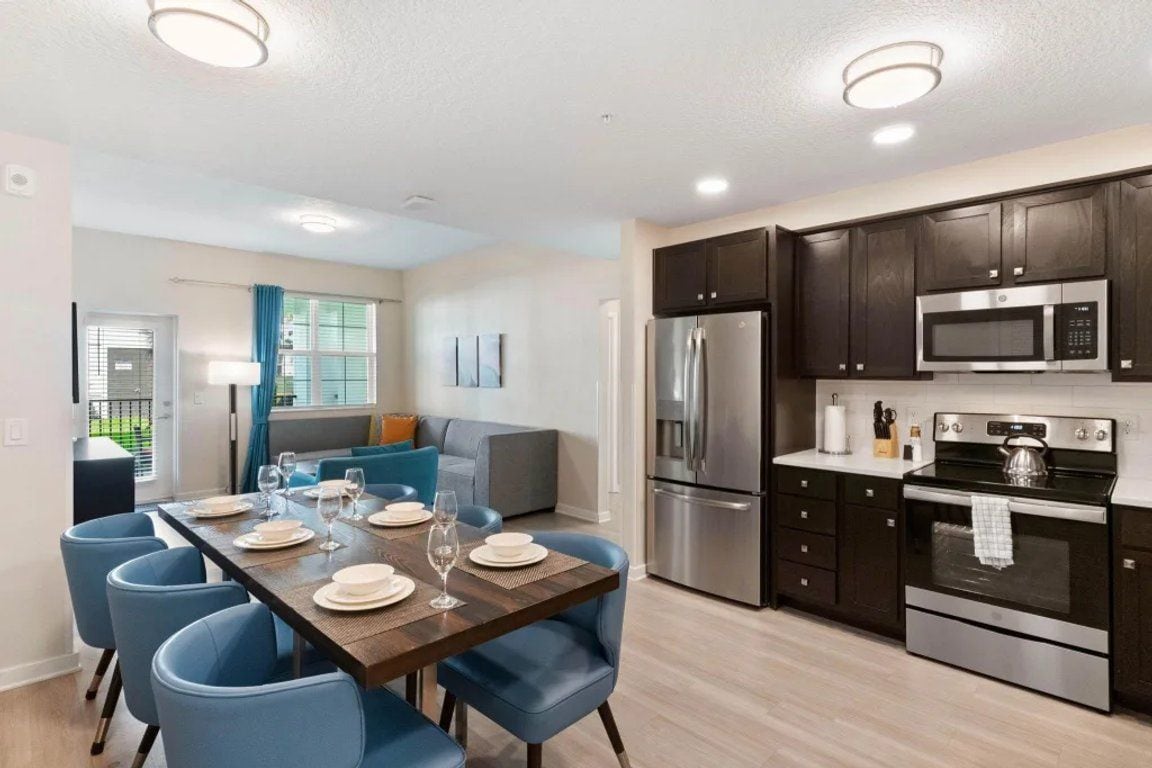 A modern kitchen with dark cabinetry, stainless steel appliances, and a dining area with blue chairs and a wooden table set for a meal.