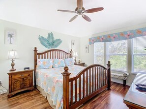 Ein Schlafzimmer mit einem hölzernen Bett, einem Deckenventilator, einem Nachttisch, einer Lampe und einem Fenster mit Jalousien.