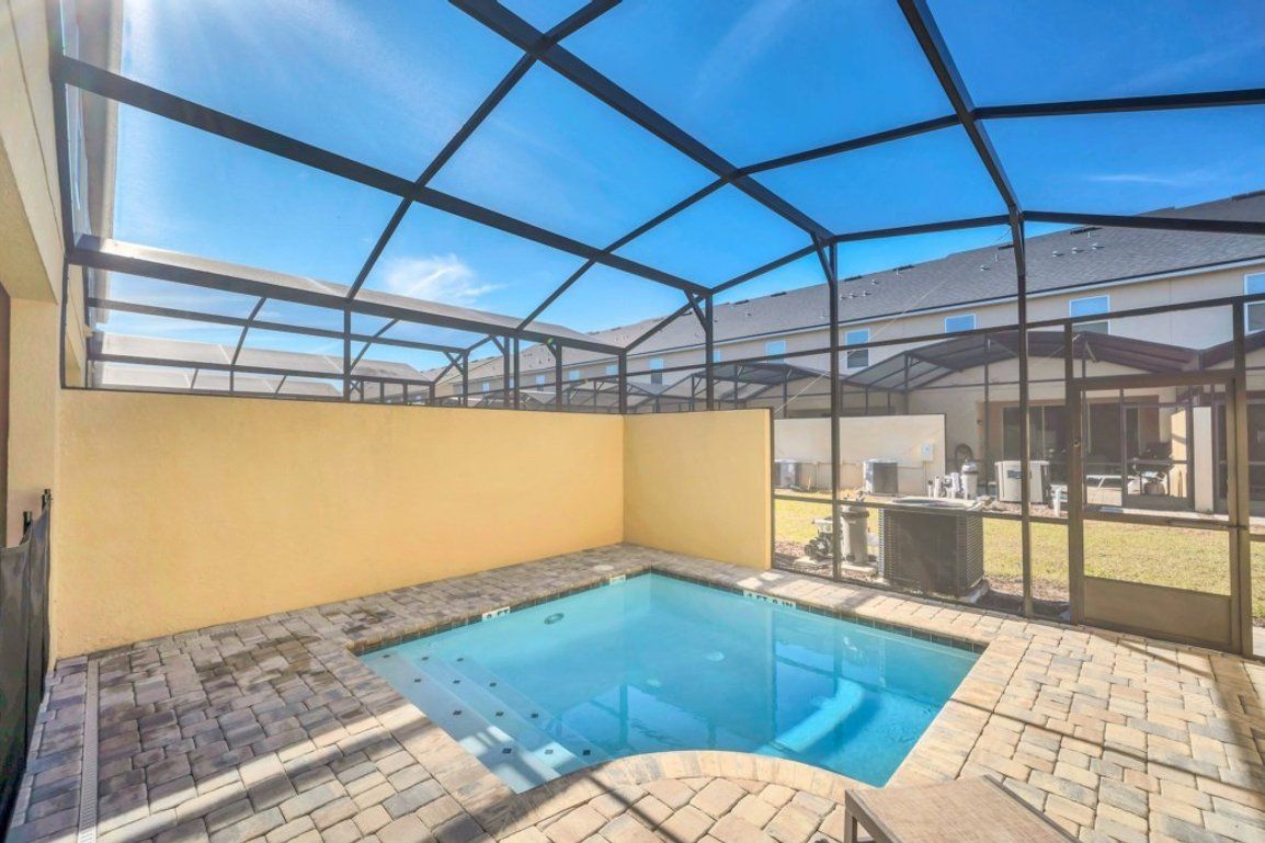 A covered outdoor swimming pool with a clear roof structure, a brick patio, and a view of a building in the background.