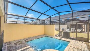 A covered outdoor swimming pool with a clear roof structure, a brick patio, and a view of a building in the background.