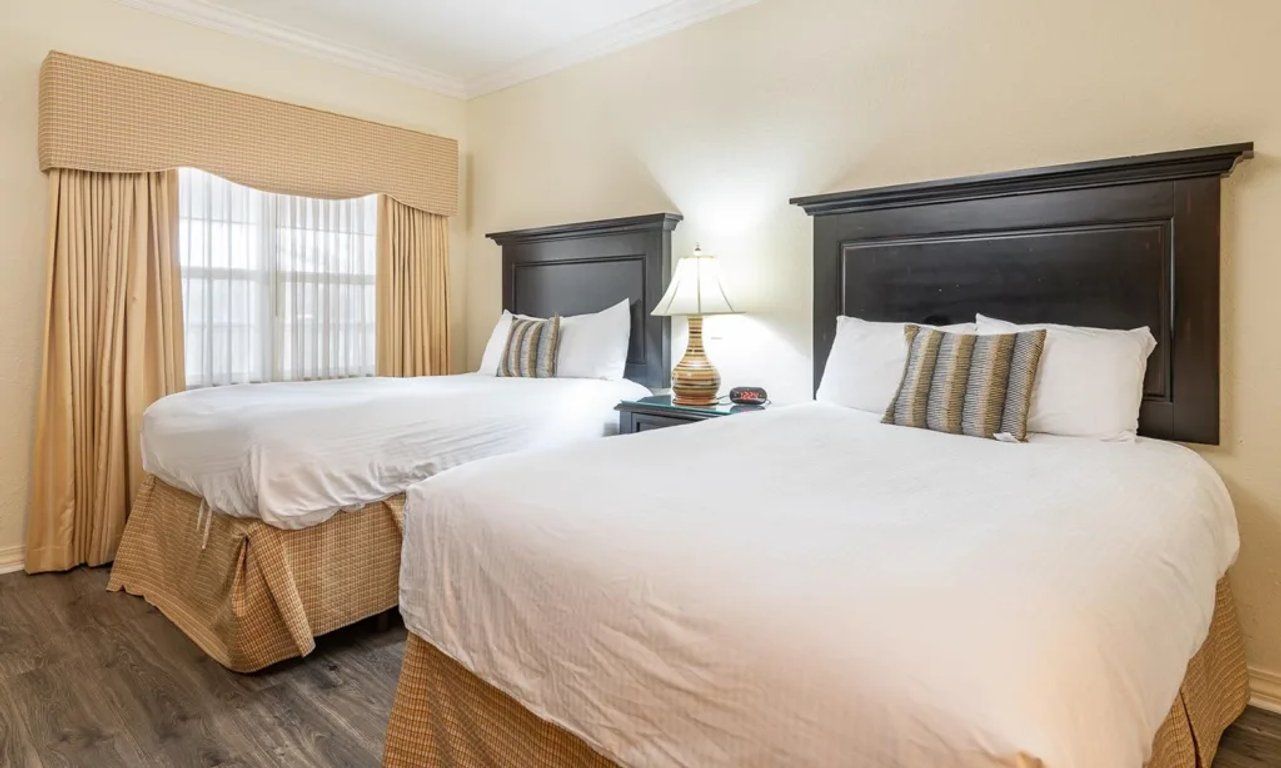 A hotel room with two beds, a nightstand with a lamp, and a window with curtains.