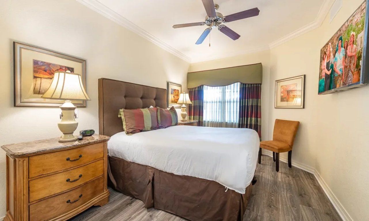 Une chambre d’hôtel avec un lit, une table de chevet, une chaise, un ventilateur de plafond et des œuvres d’art accrochées aux murs.