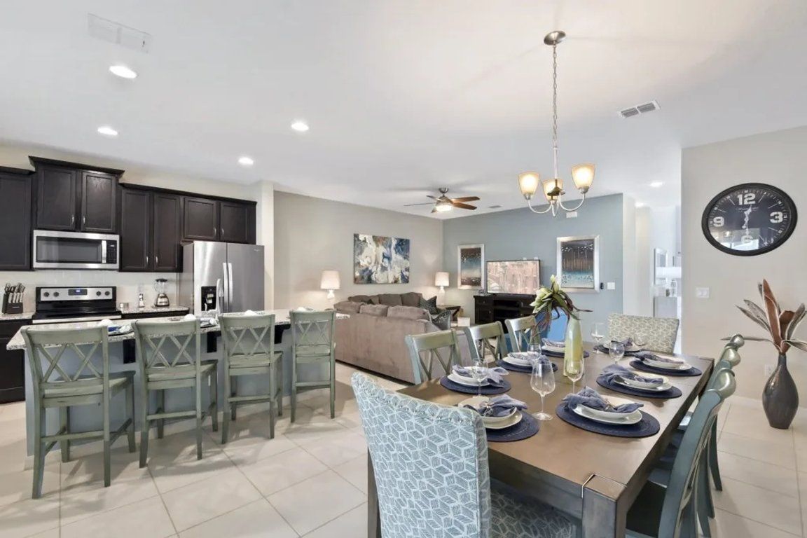 A modern kitchen with dark cabinets, a microwave, and a refrigerator. There is a dining area with a table set for four, chairs, and a chandelier.