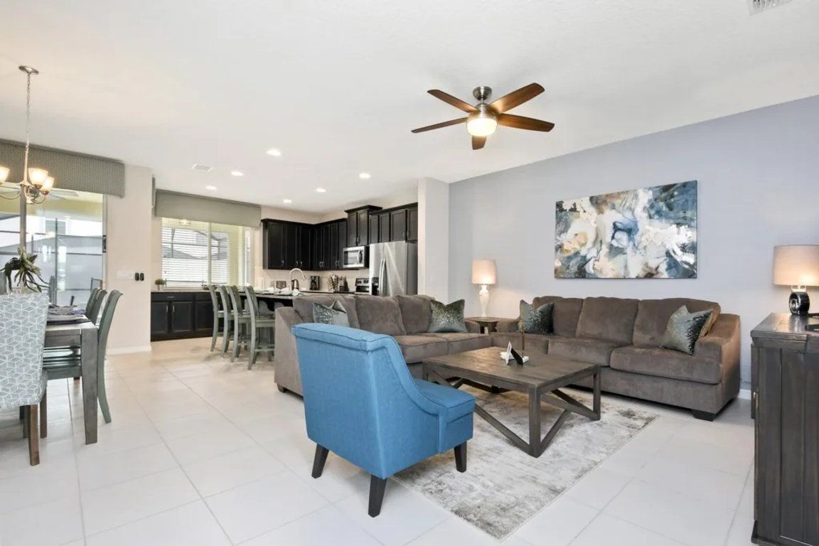 A modern living room with a blue armchair, a gray sofa, and a coffee table. There is a ceiling fan, a painting on the wall, and a dining area with a table and chairs.