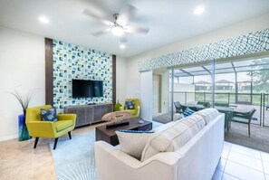 A modern living room with a flat-screen TV, a sofa, and a coffee table. There is a patterned wall and a glass door leading to an outdoor area. - Windsor at Westside - 4763 (Kissimmee)