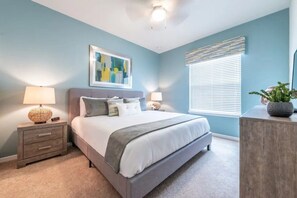 A bedroom with a bed, nightstands, a lamp, a painting, and a window with blinds. - Windsor at Westside - 4763 (Kissimmee)