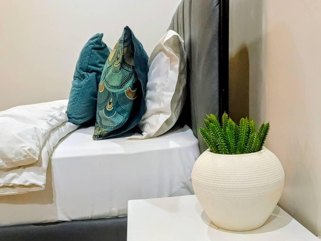 Een bed met wit beddengoed, een blauwgroen kussen, een wit kussen en een groene plant in een pot met structuur.
