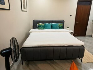 A bedroom with a bed, a bedside table, a fan, and a door. - Luxury studio kyanja ring road (Kampala)