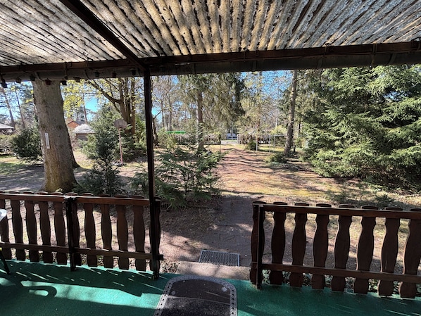 A covered outdoor area with a wooden railing, a green mat, and a view of a forested path.
