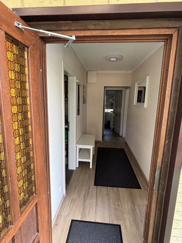 A hallway with a wooden door, a white bench, a black mat, and a view into another room.