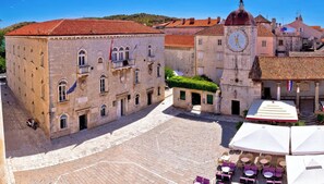 Um edifício histórico de pedra com uma torre do relógio, um pátio pavimentado e área de estar ao ar livre com guarda-sóis.