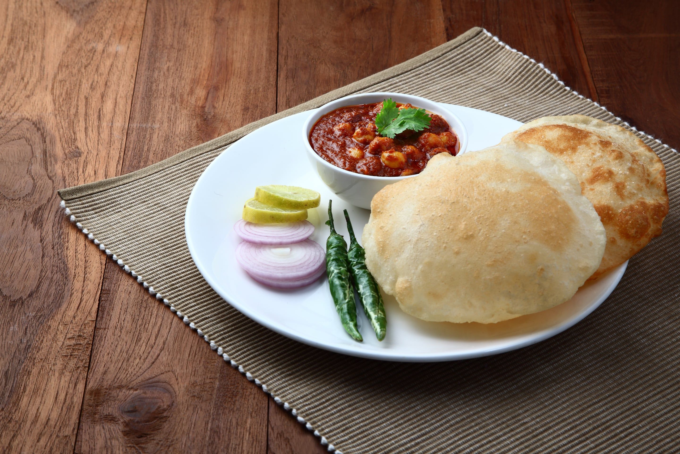 A plate with a bowl of curry, two pieces of bread, slices of onion, a lemon wedge, and two green chilies.