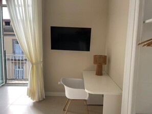 Una habitación con una silla blanca, un televisor de pantalla plana montado en la pared y una mesita con una lámpara.
