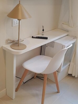 Un escritorio blanco con una lámpara, un control remoto y una silla blanca en una habitación con un radiador blanco y cortinas.