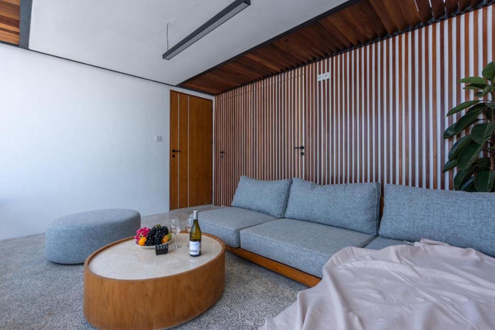 A modern living room with a grey sectional sofa, a round wooden coffee table with a bottle and fruit, and a potted plant.