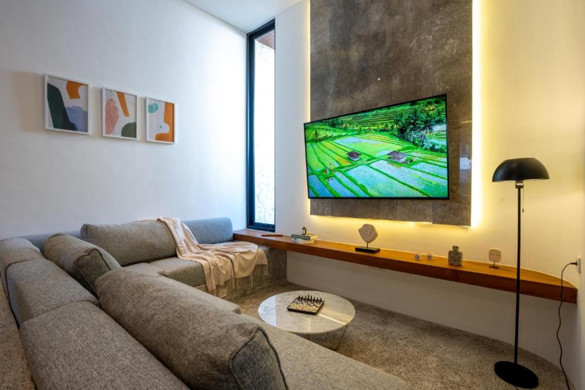 A modern living room with a large flat-screen TV mounted on the wall, a grey sectional sofa, a small round coffee table, and a floor lamp.