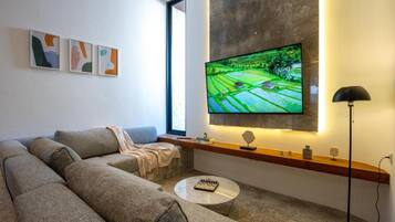 A modern living room with a large flat-screen TV mounted on the wall, a grey sectional sofa, a small round coffee table, and a floor lamp.