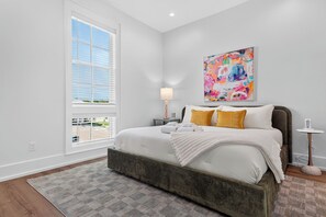A bedroom with a large bed, a bedside table with a lamp, a colorful abstract painting on the wall, and a view through the window. - SeaLuxe Brand New Construction, Beach Front, Elevated Pool, Hot Tub,  Sleeps 14 (St. George Island)
