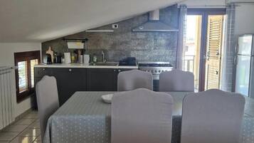 A dining area with a table and chairs, a kitchen in the background, and a view through an open door.