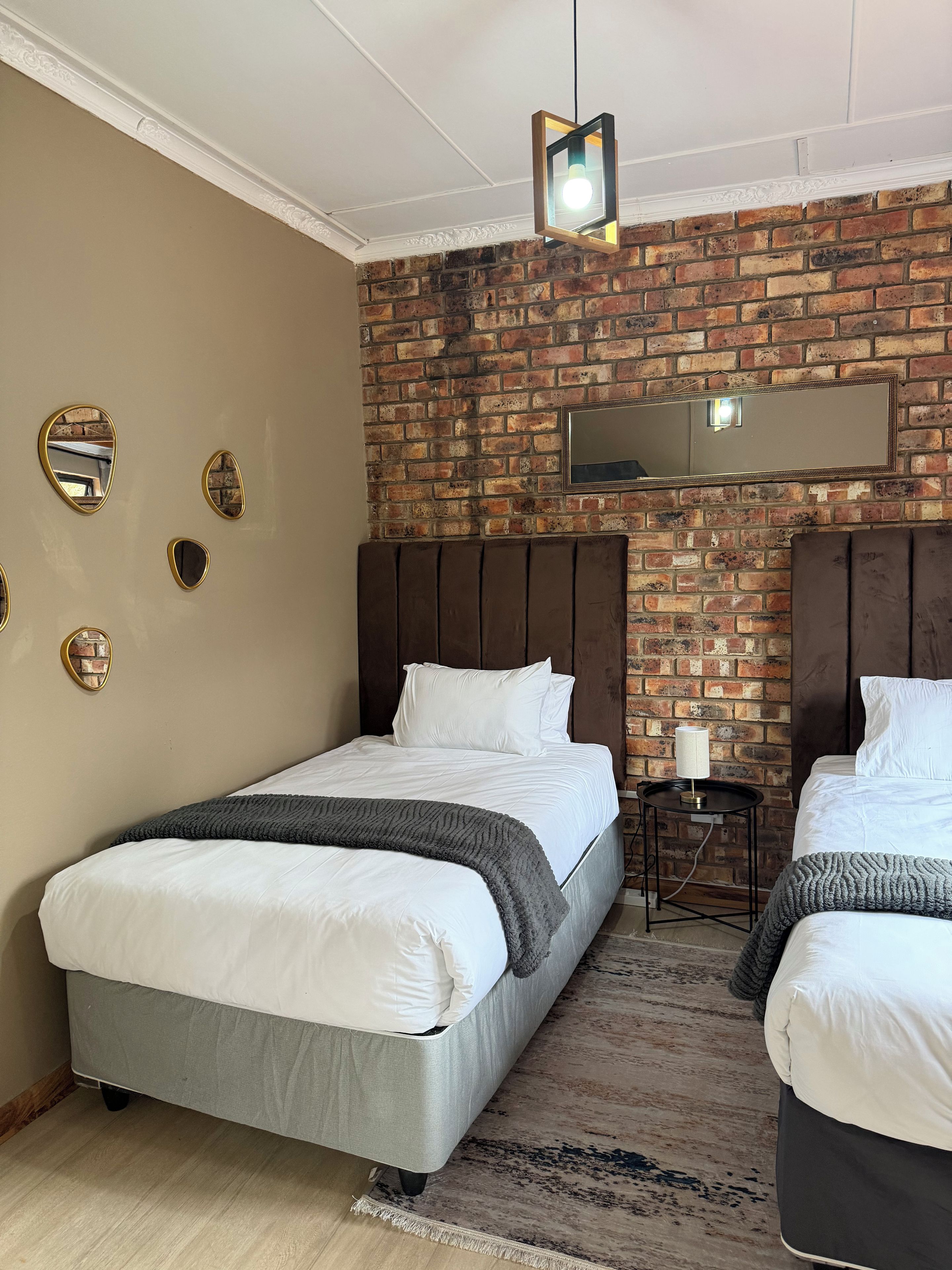 Een hotelkamer met een bakstenen muur, twee bedden met wit beddengoed, een kleine tafel met een lamp en een plafondlamp.