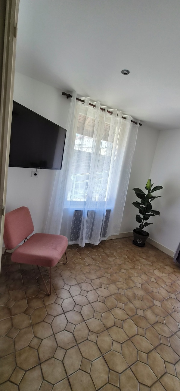 Habitació amb una cadira rosa, una planta en test i una televisió penjada a la paret, i una finestra amb un teixit translúcid.