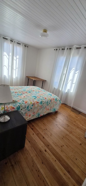 Un dormitori amb terra de fusta, un llit amb llençol floral, una taula de nit amb una làmpada i una finestra amb cortines.