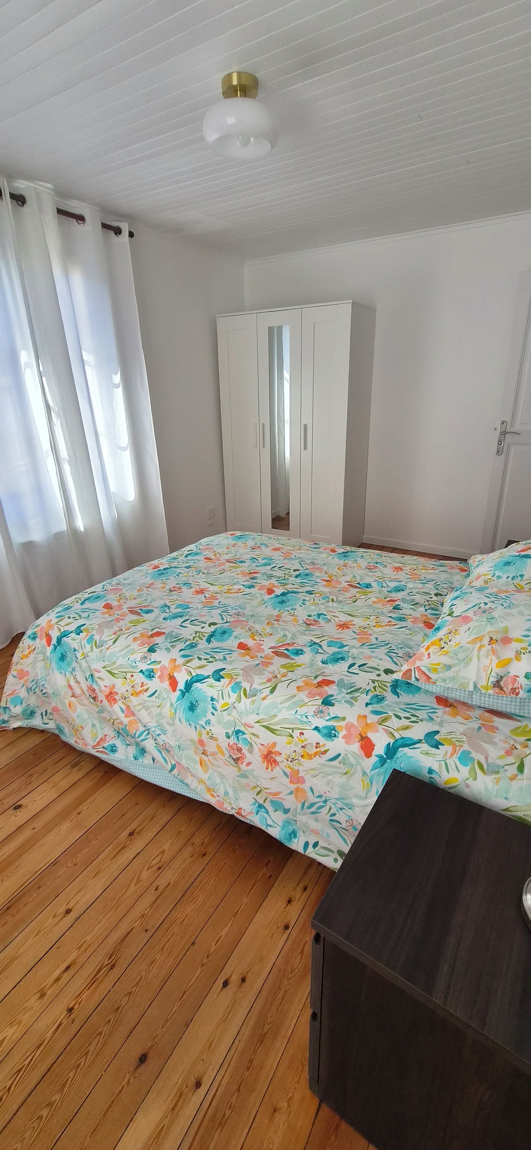 Un dormitorio con piso de madera, una cama con colcha estampada, cortinas blancas y un armario.