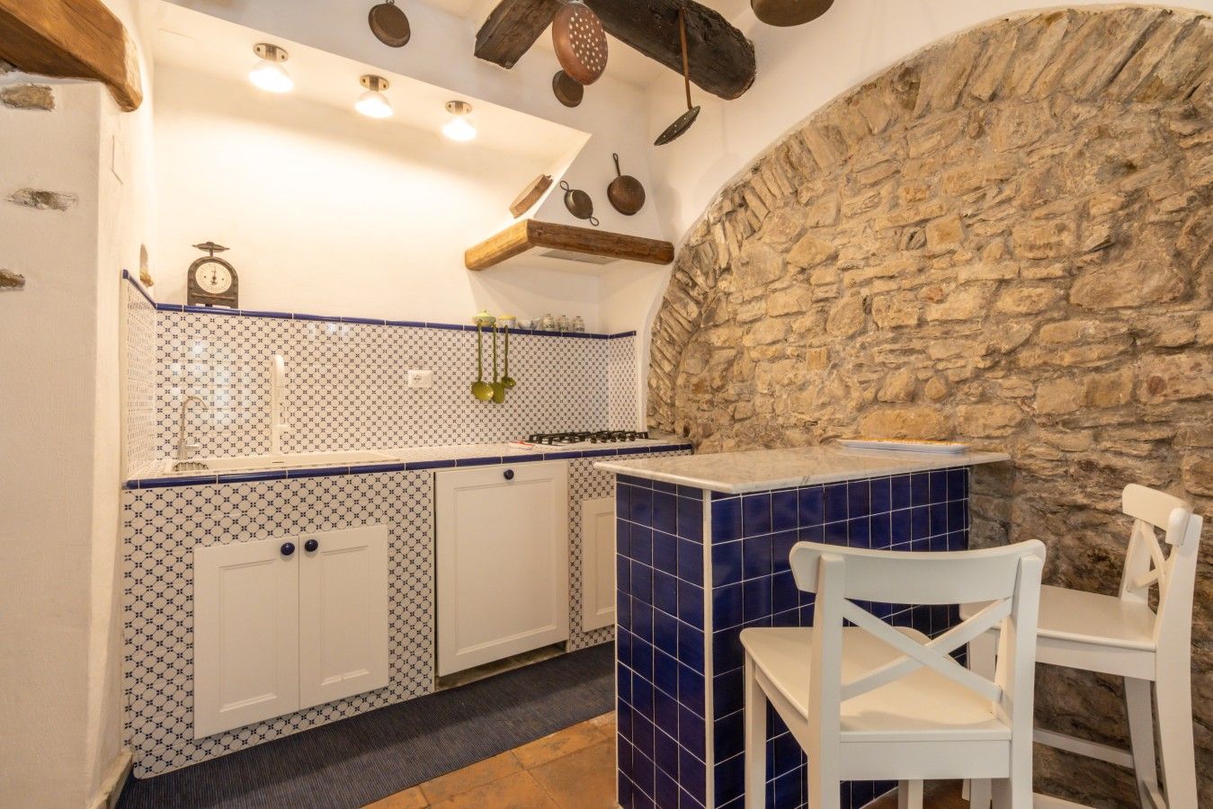A kitchen with white cabinets, blue patterned backsplash, stone wall, hanging utensils, and a white chair.