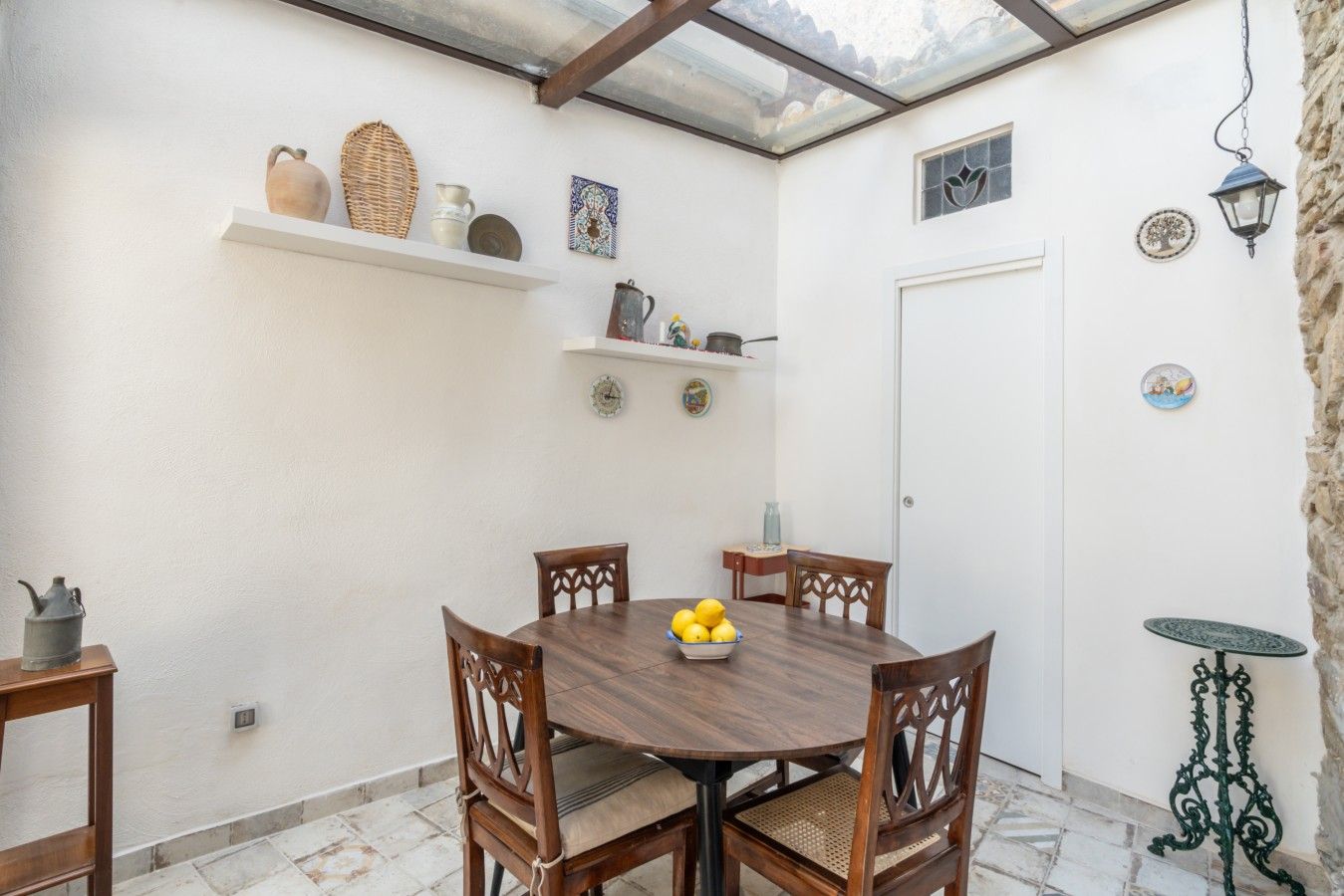 A patio with a wooden table and chairs, a small shelf with decorative items, and a door.