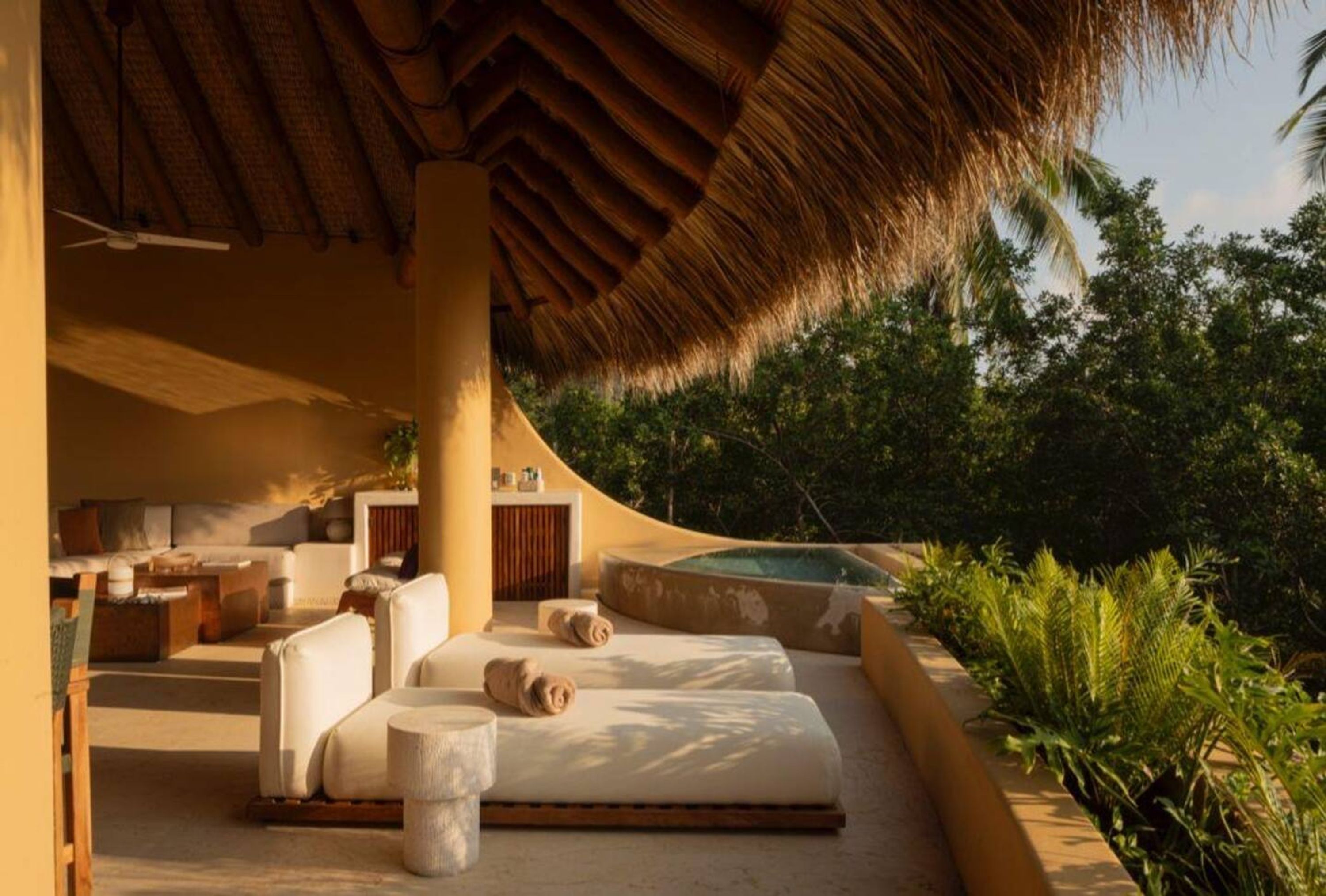 Un área de descanso al aire libre con luz solar, un arreglo de asientos curvos, un techo de paja y una piscina.