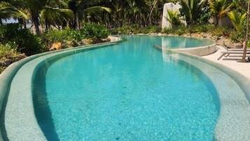 Una piscina azul cristalina con borde curvo, rodeada de palmeras y follaje.