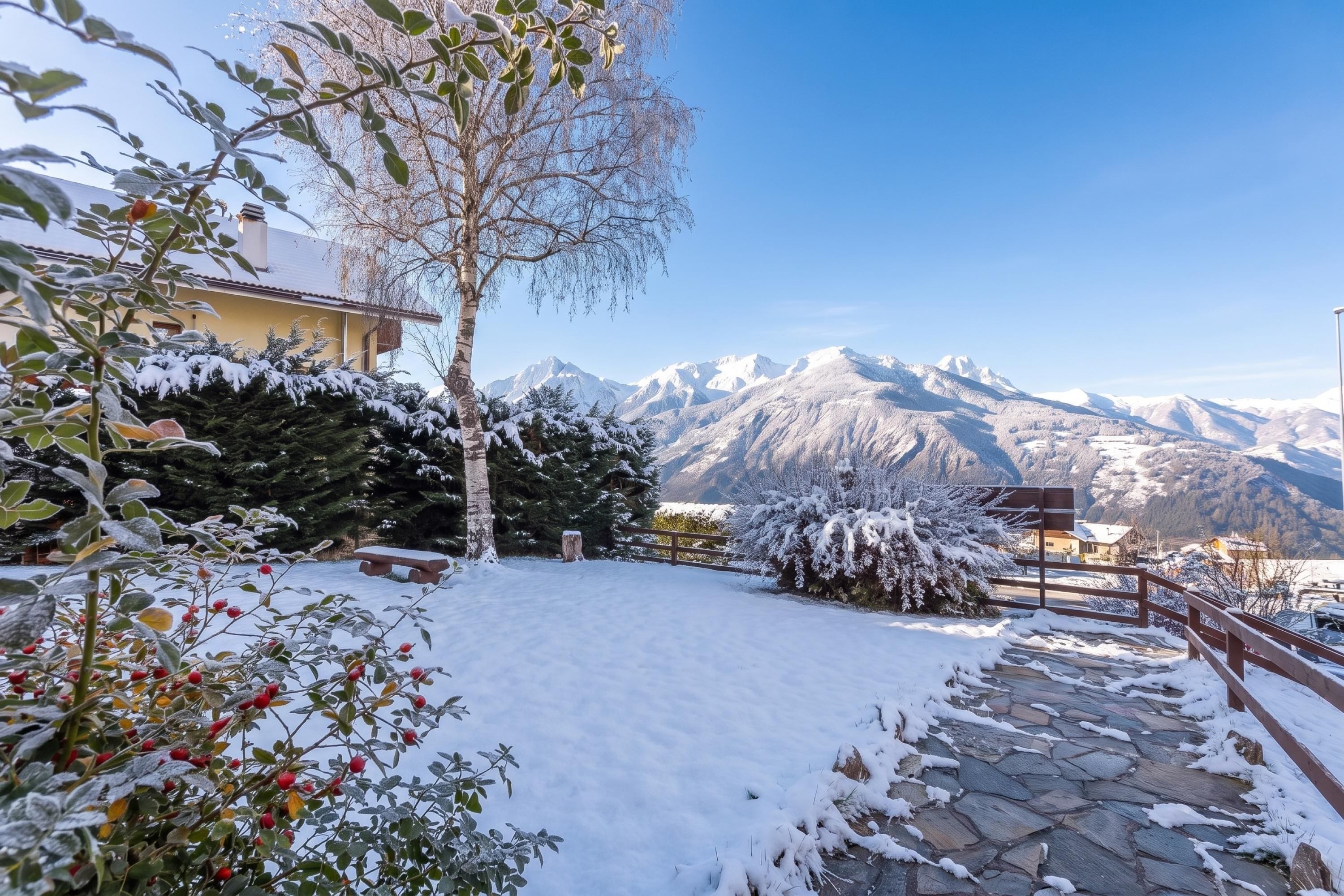 A snow-covered garden with a stone pathway leading to a bench, surrounded by trees and a mountain view.