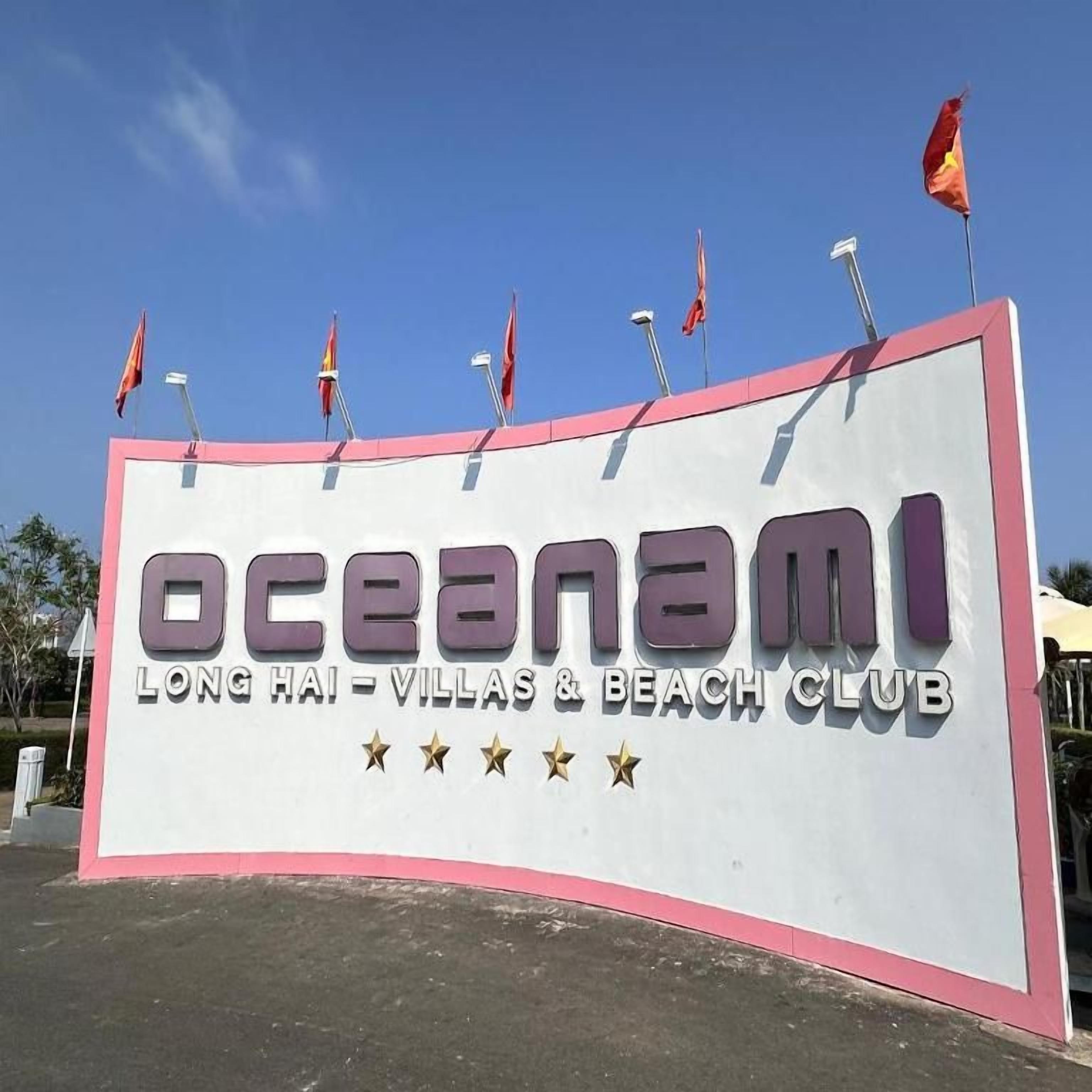 A large sign for Oceanami Long Hai - Villas & Beach Club with multiple flags on top.