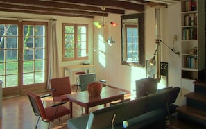 Een kamer met een houten plafond, een tafel, stoelen, een boekenkast en grote ramen.