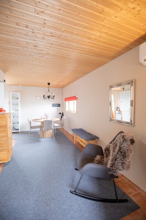 A room with a wooden ceiling, a dining table with chairs, a bench, a fluffy chair, a mirror on the wall, and a white cabinet. - Geilin 11 (Innan Glyvur)