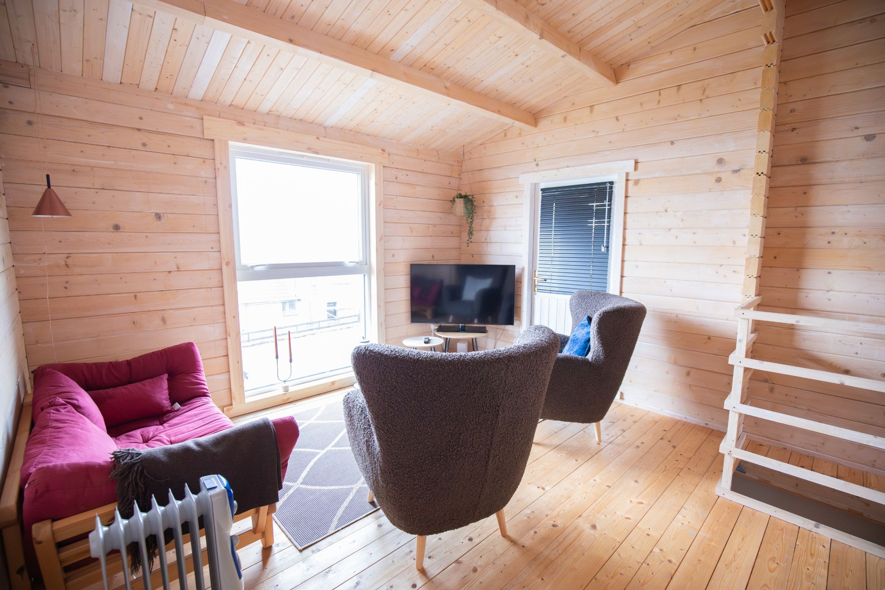 A cozy cabin interior with a wooden floor, a sofa, a chair, a TV, and a window with blinds.