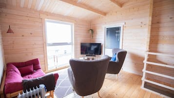 A cozy cabin interior with a wooden floor, a sofa, a chair, a TV, and a window with blinds.