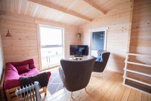 A cozy cabin interior with a wooden floor, a sofa, a chair, a TV, and a window with blinds. - Geilin 11 (Innan Glyvur)