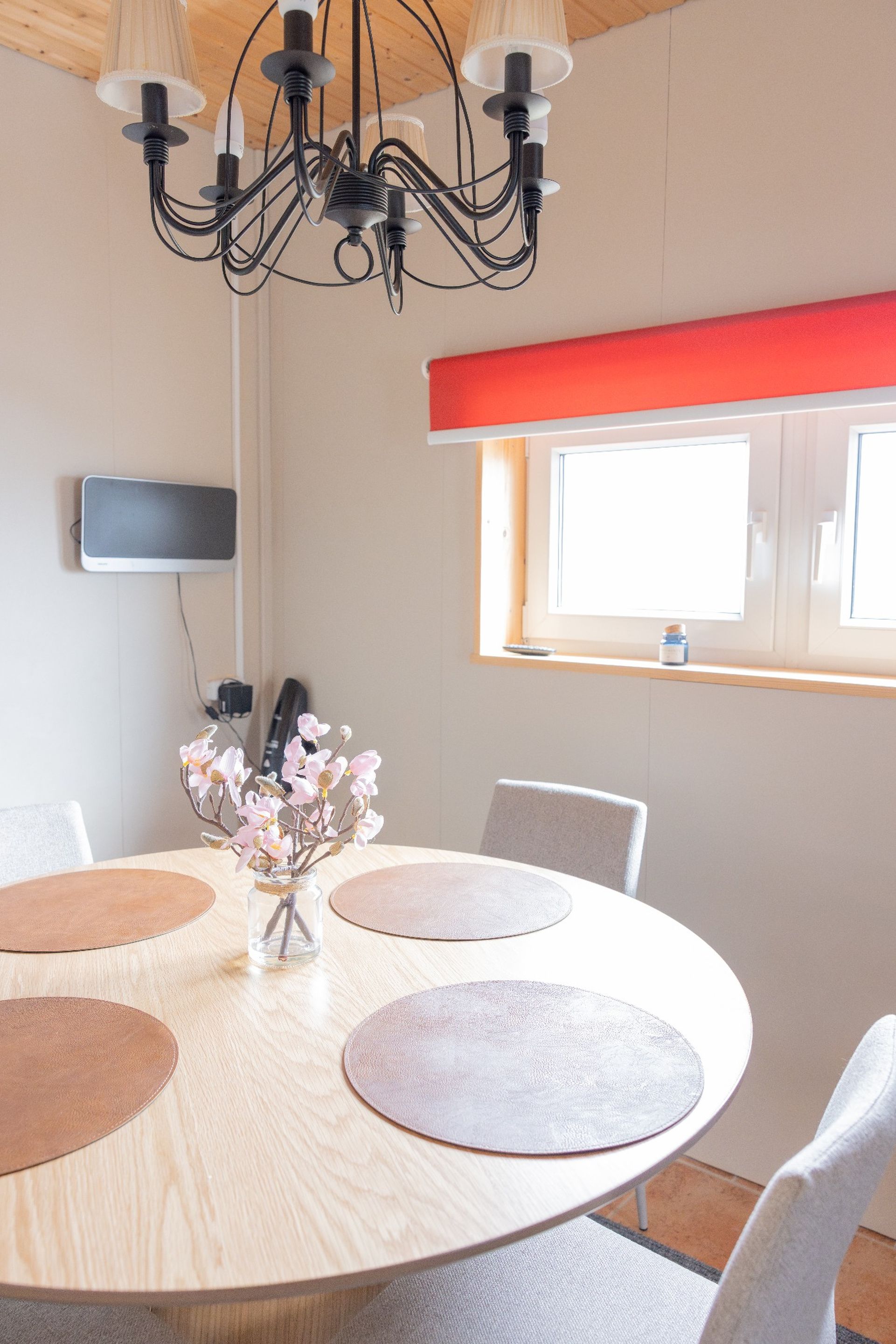 A modern dining area with a round table, chairs, a chandelier, a vase with flowers, and a window with blinds.
