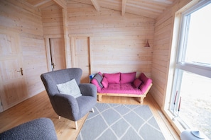 A cozy room with wooden walls, a grey armchair, a pink sofa, and a window. - Geilin 11 (Innan Glyvur)